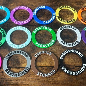 Puede incluir: Una colección de fichas de juego circulares y coloridas con etiquetas de efectos de estado como "Cegado", "Hechizado" y "Agotado". Las fichas son de color azul, morado, verde y naranja, con letras blancas. Están dispuestas sobre una superficie de madera oscura.