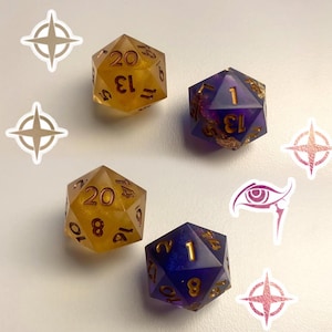May include: Four twenty-sided dice arranged in a square pattern. Two dice are translucent amber with gold numbering, and two are translucent purple with gold numbering. Decorative stickers of stars and an eye are placed around the dice.