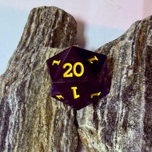 May include: A twenty-sided die featuring a deep purple translucent finish with glitter accents and bright yellow engraved numbers. The die displays the number 20 prominently on its top face, with other numerical values visible on the surrounding facets.