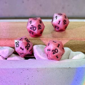 May include: Four twenty-sided dice with a pearlescent pink finish and black ink numbering. Each die features a distinct geometric shape with multiple triangular faces, displaying numbers including 1, 13, 19, and 20 clearly etched on the surface.