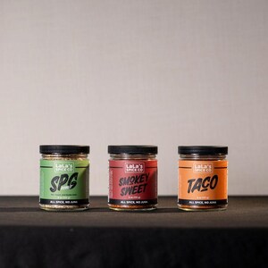 May include: Three glass spice jars with black lids are displayed on a black surface. The jars have labels with the text "SPG", "Smokey Sweet", and "Taco". The labels are green, red, and orange, respectively, and include the text "Lala's Spice Co."