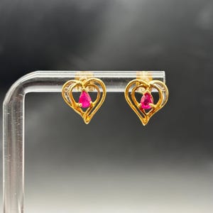 May include: A pair of gold-colored heart-shaped stud earrings. Each earring features a teardrop-shaped, bright pink gemstone set within the heart's center. The hearts are outlined with small, clear stones.