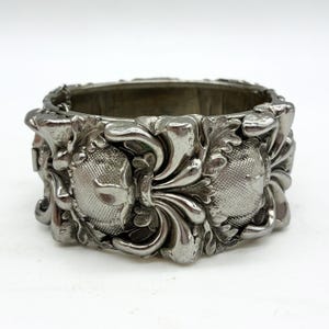May include: A silver-toned metal hinged cuff bracelet featuring a prominent embossed floral and leaf motif. The design includes textured, rounded central elements surrounded by intricate, raised botanical patterns with a polished finish.
