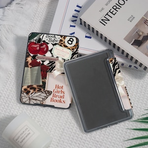 May include: A black phone case with a collage design featuring cherries, leopard print, a butterfly, and the text "Hot Girls Read Books." A clear phone case sits next to it. Books and a candle are in the background.