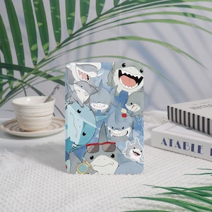 Pode incluir: Capa de telemóvel retangular com um divertido padrão de tubarões. O design apresenta vários tubarões de desenho animado em tons de cinza e azul, alguns usando óculos de sol e sorrindo. O fundo é azul claro com detalhes brancos.