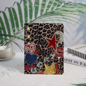 Può includere: Una copertina di diario con stampa leopardata e varie decorazioni. Include una stella rossa, un cuore di jeans, una stella dorata e la scritta "ALL YOU NEED IS LOVE". Sono presenti anche una tigre bianca e labbra rosse.