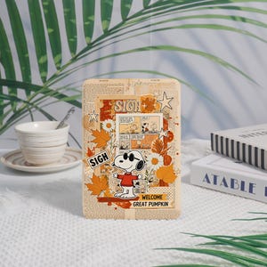 May include: A rectangular wooden plaque featuring a Snoopy cartoon design with the text "WELCOME GREAT PUMPKIN". The design includes fall leaves, daisies, and comic strip panels. The plaque has a vintage newspaper background.