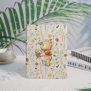 May include: A rectangular tablet case featuring a cream background with a repeating pattern of small yellow, orange, and blue wildflowers. In the center, a yellow bear character holds a honey pot labeled HUNNY with a small bee hovering above.