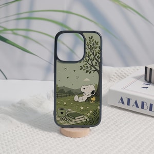 May include: A dark-rimmed phone case featuring a printed illustration of Snoopy and Woodstock relaxing under a tree in a green meadow. The scene includes a picnic blanket, a coffee mug with a heart, and a book, all set against a soft green sky.