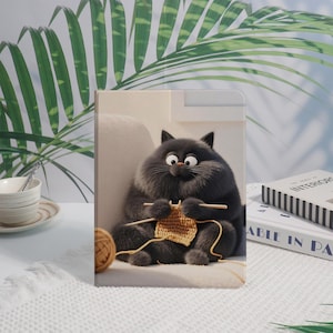 Puede incluir: Una funda rectangular para tableta con una ilustración gráfica de un gato negro redondo y esponjoso sentado, tejiendo con lana de color marrón claro. El gato tiene ojos grandes y sostiene dos agujas de tejer de madera mientras trabaja.