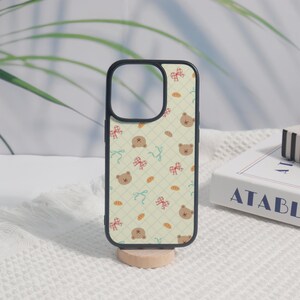 May include: A black-framed phone case with a light green background featuring a repeating pattern of brown teddy bear faces, red bows, and bread shapes. The case is displayed on a small wooden stand.