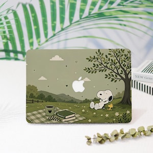 May include: A sage green laptop hard shell case featuring a printed illustration of Snoopy and Woodstock relaxing under a tree in a grassy field. The design includes a picnic blanket, a coffee mug, books, and a white Apple logo cutout in the center.