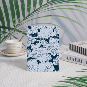 May include: A rectangular notebook with a blue and white cloud pattern. The design features swirling clouds against a dark blue background. The notebook is displayed on a white surface, with a cup and saucer and a book in the background.