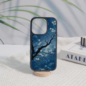 May include: A phone case with a black frame and a blue and white floral design. The case features a dark blue background with a tree and white flowers. The case is displayed on a small wooden stand.