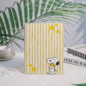Op de afbeelding: Een notitieboek met een geel en wit gestreept patroon en gele sterren. De kaft toont een cartoon Snoopy-personage dat een gele vogel omhelst. Het notitieboek ligt op een wit oppervlak.
