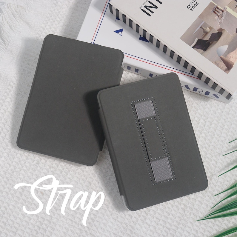 May include: Two gray tablet cases are displayed, one open and one closed. The open case has a gray strap for support. The word "Strap" is written in cursive across the bottom of the image. A book is in the background.
