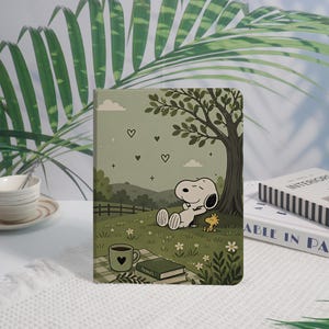 Può includere: Una custodia rettangolare per tablet con un'illustrazione color verde salvia di Snoopy e Woodstock che si rilassano sotto un albero in un prato. La scena include una tazza di caffè, libri, piccoli fiori bianchi, un recinto e cuori.
