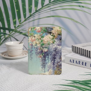 Può includere: Una custodia rettangolare con un dipinto floreale di glicine viola a cascata, fiori rosa e bianchi e un cielo azzurro. La custodia è appoggiata su una superficie bianca, con una tazza da tè e libri sullo sfondo.