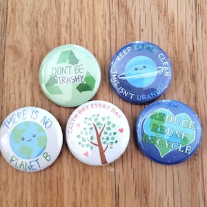 May include: Five round, colorful buttons with environmental themes. One button says "Don't Be Trashy" with a recycling symbol. Another says "Keep Earth Clean, This Isn't Uranus". Other buttons say "There Is No Planet B", "Earth Day Every Day", and "Reduce, Reuse, Recycle".