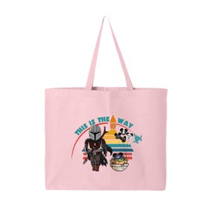 May include: A pale pink tote bag with a cartoon illustration of The Mandalorian and Grogu. The design includes the text "THIS IS THE WAY" and colourful accents. The bag has a rectangular shape and two handles.
