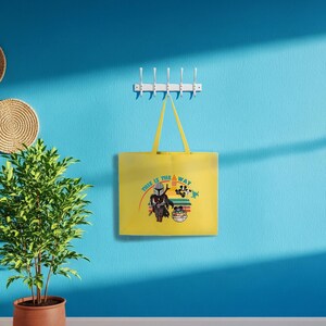 May include: A yellow tote bag featuring the phrase "THIS IS THE WAY" and a graphic of characters from a popular science fiction series. The bag hangs on a white hook rack against a blue wall. A potted plant and wall decorations are also visible.