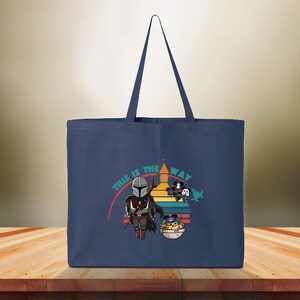 May include: Navy blue tote bag with a graphic of The Mandalorian and Grogu, and the text "THIS IS THE WAY". The design includes a colorful sunset-like background. The bag has a simple, rectangular shape with a long handle.