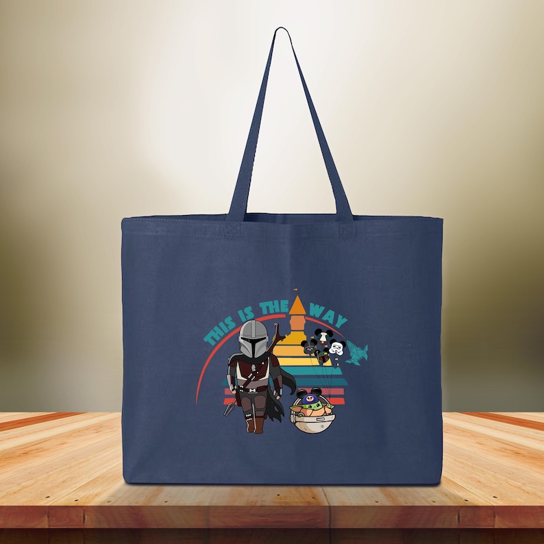 May include: Navy blue tote bag with a graphic of The Mandalorian and Grogu, and the text "THIS IS THE WAY". The design includes a colorful sunset-like background. The bag has a simple, rectangular shape with a long handle.
