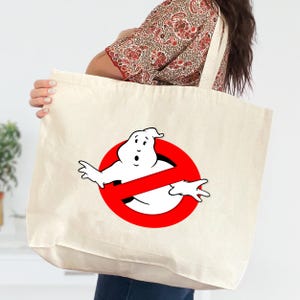 May include: Cream-colored tote bag featuring the Ghostbusters logo: a white ghost inside a red circle with a diagonal red line. The bag has long, cream-colored straps and is held by a person.