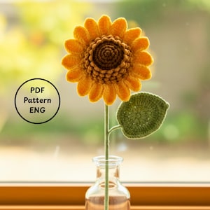 May include: A crocheted sunflower with yellow petals and a brown center, green stem and leaf, and a glass vase. The image includes a circular graphic with the text "PDF Pattern ENG".