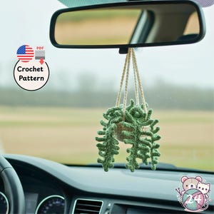 Könnte beinhalten: Eine gehäkelte Hängepflanze, die am Rückspiegel eines Autos befestigt ist. Die Pflanze hat grüne, strukturierte Blätter und einen beigen Topf. Das Bild enthält eine "Crochet Pattern"-Grafik mit einer amerikanischen Flagge.