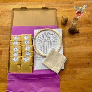 Puede incluir: Un kit de manualidades en una caja de cartón abierta, que contiene suministros de bordado. Incluye un bastidor de bordado de madera con un diseño floral, tela y varios paquetes de papel marrón. El kit está dispuesto sobre papel de seda morado.