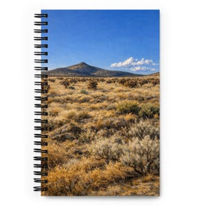 May include: Spiral-bound notebook featuring a landscape image. The cover displays a scenic view of a mountain under a bright blue sky with a few clouds. The foreground shows dry, golden grasses and shrubs.