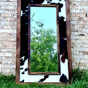 May include: Rectangular mirror with a rustic wooden frame. The frame is accented with cowhide in black and white patterns. The mirror reflects a green tree and blue sky.
