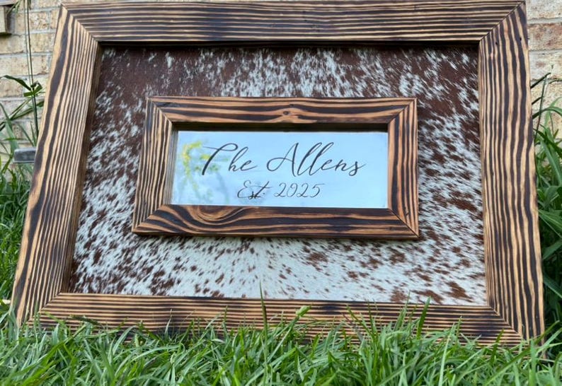 Pu&ograve; includere: Un cartello rustico con cornice in legno con sfondo in pelle di mucca. Il cartello ha una cornice interna pi&ugrave; piccola con il testo "The Allens Est. 2025" in elegante scrittura. Il legno ha una finitura scura e bruciata.