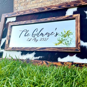 Pu&ograve; includere: Specchio rettangolare con cornice in legno e sfondo in pelle di mucca. Lo specchio riporta la scritta "The Gilmore's Est. May 2027" in corsivo nero. La cornice &egrave; in legno scuro con venature naturali.