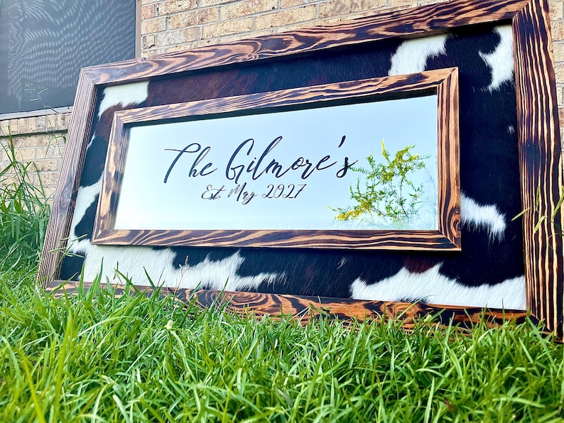 Pu&ograve; includere: Specchio rettangolare con cornice in legno e sfondo in pelle di mucca. Lo specchio riporta la scritta "The Gilmore's Est. May 2027" in corsivo nero. La cornice &egrave; in legno scuro con venature naturali.