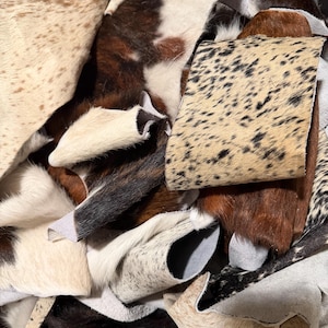 May include: A collection of assorted cowhide leather scraps featuring various natural patterns, including spotted, speckled, and solid brown, black, and white textures. The pieces have a soft fur finish and irregular shapes.