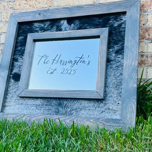 Pu&ograve; includere: Un'opera d'arte incorniciata con una cornice in legno grigio. Lo strato interno presenta un motivo a pelle di mucca e uno specchio incorniciato pi&ugrave; piccolo &egrave; centrato, che mostra il testo "The Harrington's Est. 2025". L'opera d'arte &egrave; ambientata contro un muro di mattoni.