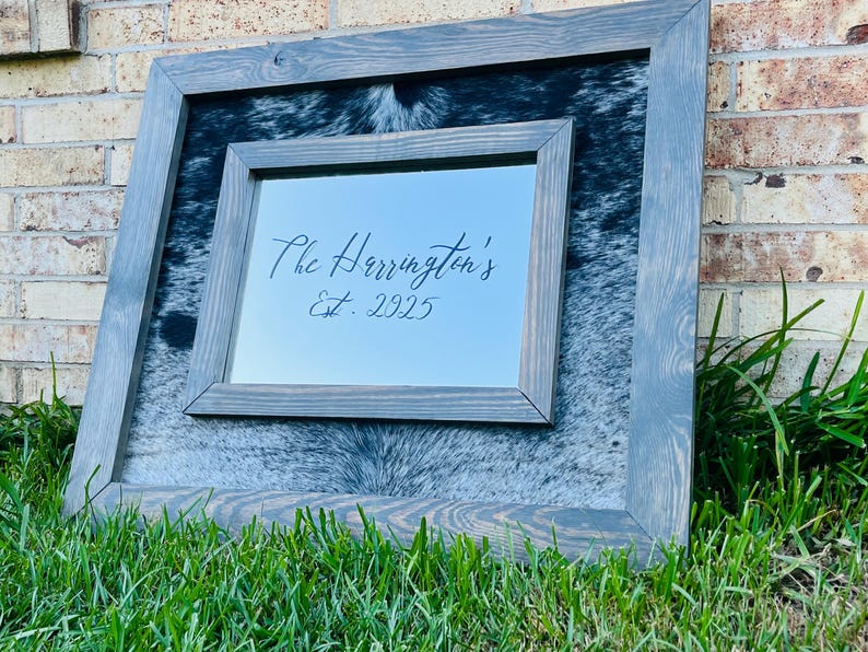 Pu&ograve; includere: Un'opera d'arte incorniciata con una cornice in legno grigio. Lo strato interno presenta un motivo a pelle di mucca e uno specchio incorniciato pi&ugrave; piccolo &egrave; centrato, che mostra il testo "The Harrington's Est. 2025". L'opera d'arte &egrave; ambientata contro un muro di mattoni.