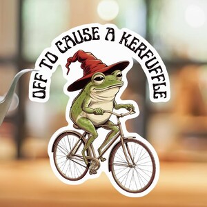 May include: A die-cut sticker featuring a green frog wearing a dark red pointed hat, riding a brown bicycle. The text "OFF TO CAUSE A KERFUFFLE" is arched above the frog in black lettering. The sticker is held by a pair of silver metal tweezers.