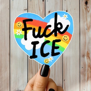 Puede incluir: Una pegatina en forma de coraz&oacute;n con las palabras "Fuck ICE" en negro, sobre un fondo de arco&iacute;ris y cielo azul. El dise&ntilde;o incluye caras sonrientes y margaritas. La pegatina se sostiene sobre un fondo de madera.