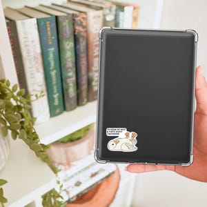 May include: A black tablet in a clear case with a sticker of a cartoon bird and the text "I'M DOING MY BEST & THAT'S DEEPLY CONCERNING". The tablet is held in a person's hand near a bookshelf with books and a plant.