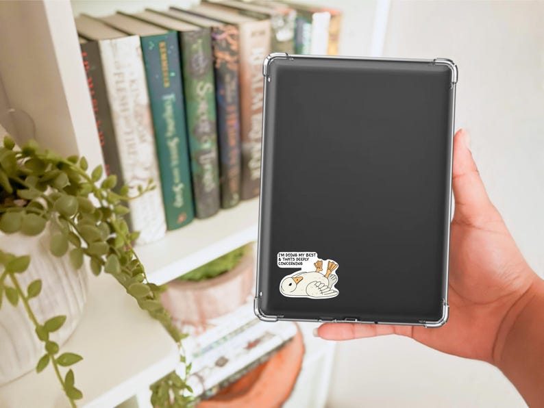 May include: A black tablet in a clear case with a sticker of a cartoon bird and the text "I'M DOING MY BEST & THAT'S DEEPLY CONCERNING". The tablet is held in a person's hand near a bookshelf with books and a plant.