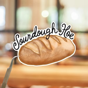 May include: A sticker featuring a golden-brown loaf of sourdough bread with a scored crust. Above the bread, the text Sourdough Hoe is written in a black, cursive script. The sticker has a white border and is being held by a pair of metal tweezers.