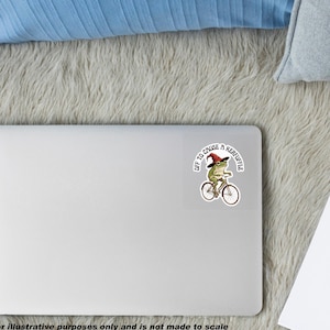 May include: A square sticker featuring a green frog wearing a red pointed hat while riding a bicycle. The text "OFF TO CAUSE A KERFUFFLE" is printed in a circular arc above the frog. The sticker is applied to the center of a silver laptop lid.