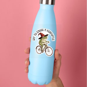 May include: A light blue metal water bottle featuring a sticker of a green frog wearing a red pointed hat while riding a bicycle. The sticker includes the text "OFF TO CAUSE A KERFUFFLE" arched above the frog in a bold, black, sans-serif font.