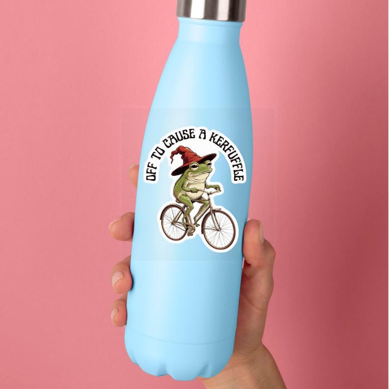 May include: A light blue metal water bottle featuring a sticker of a green frog wearing a red pointed hat while riding a bicycle. The sticker includes the text "OFF TO CAUSE A KERFUFFLE" arched above the frog in a bold, black, sans-serif font.