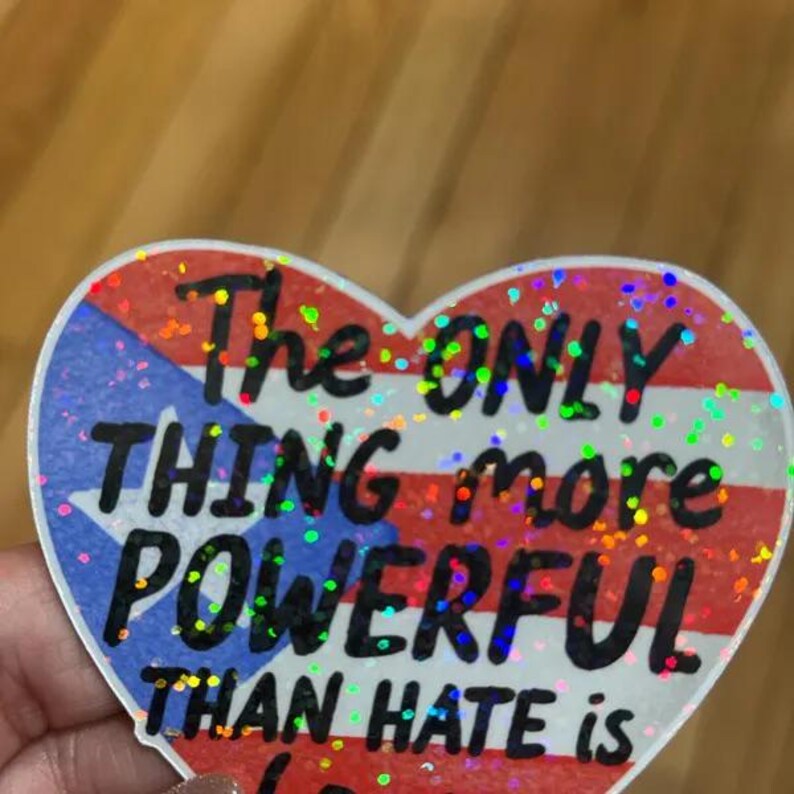 May include: Heart-shaped sticker featuring the Puerto Rican flag and the words "The ONLY THING more POWERFUL THAN HATE is LOVE". The sticker has a glittery finish.