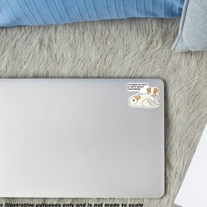 May include: A white laptop with a sticker featuring a cartoon owl lying on its back with the text "I'M DOING MY BEST & THAT'S DEEPLY CONCERNING". The laptop is on a gray, fluffy surface, with a blue pillow and a notepad.