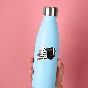 Op de afbeelding: Een lichtblauwe waterfles met een zilverkleurige metalen dop. Een sticker op de fles toont een zwarte cartoonkat met grote gele ogen en de tekst: IT'S A BLACK CAT THING. YOU WOULDN'T UNDERSTAND, vergezeld door drie kleine zwarte hartjes.